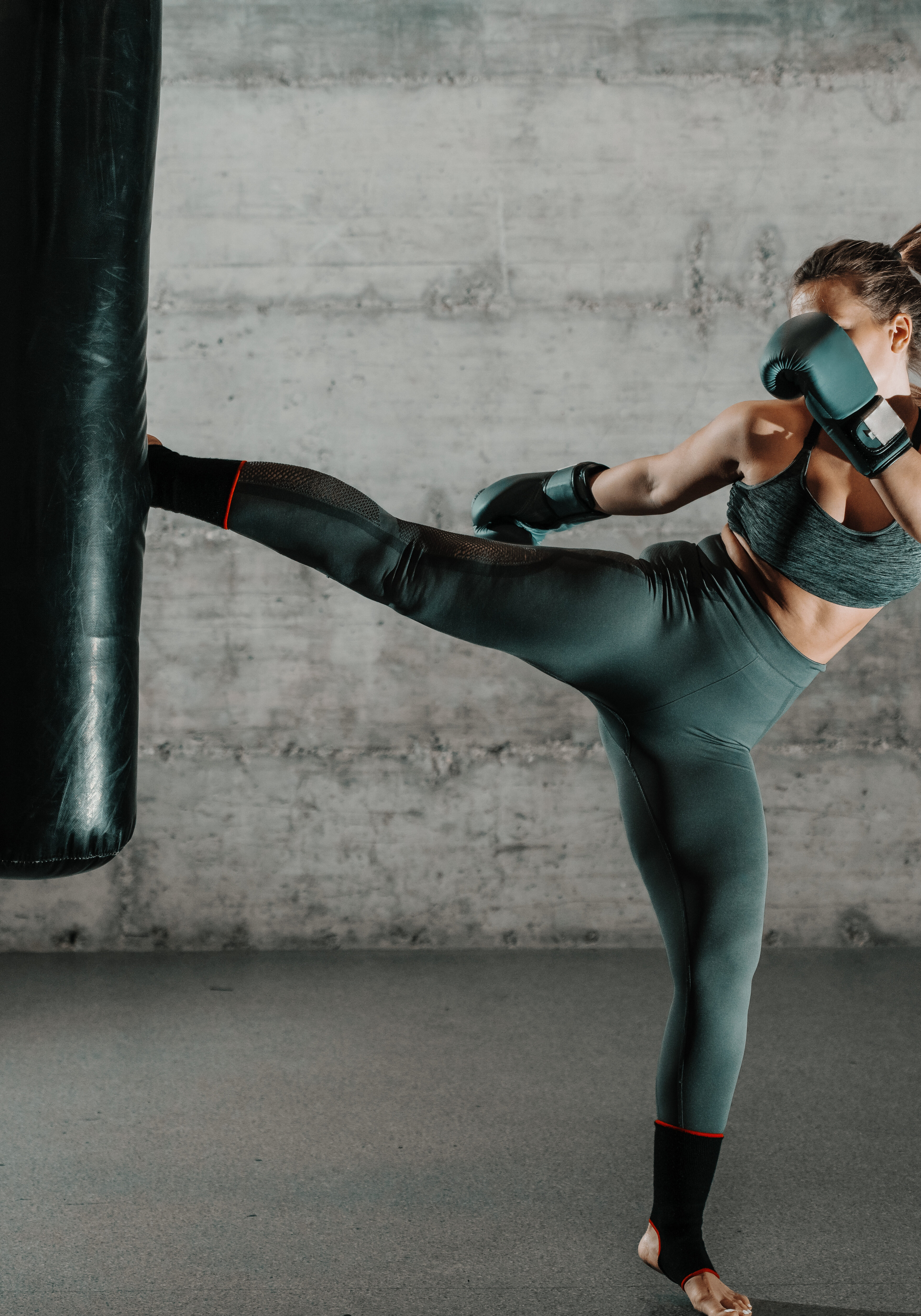 Caucasian woman in sportswear and with boxing gloves kicking bag in the gym. Full length. Wall in background.